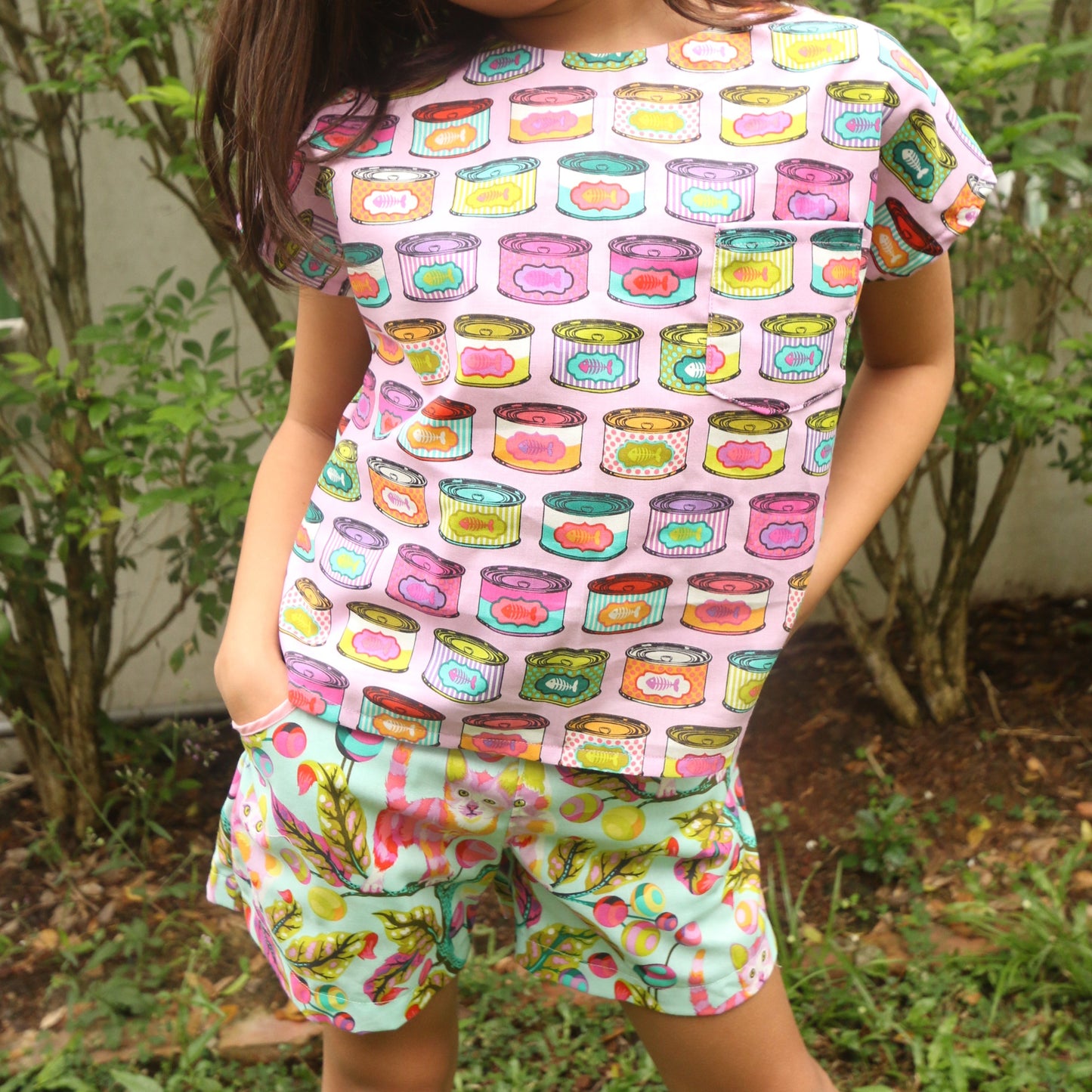 Child wearing a colorful top and shorts with a pattern of cartoonish food items and cats, standing outdoors.