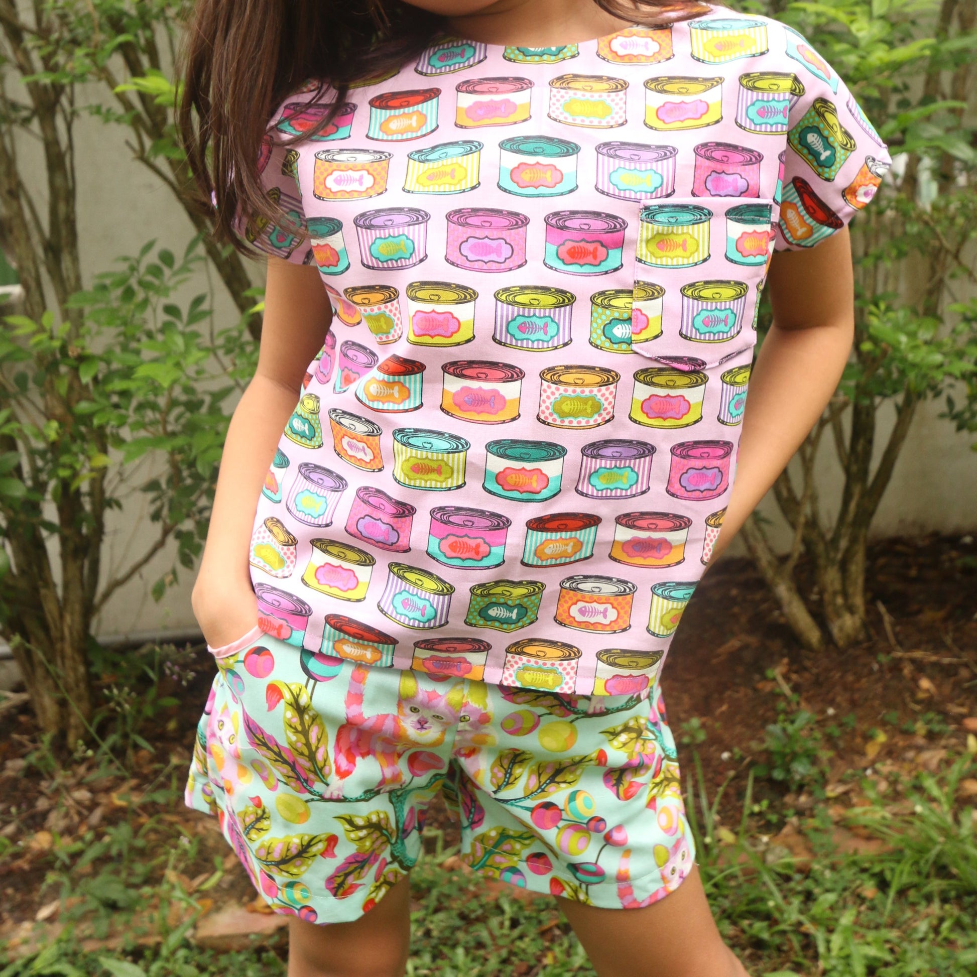 Child wearing a colorful top and shorts with a pattern of cartoonish food items and cats, standing outdoors.