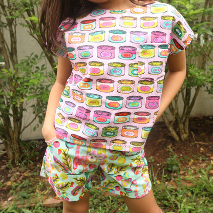 Child wearing a colorful top and shorts with a pattern of cartoonish food items and cats, standing outdoors.